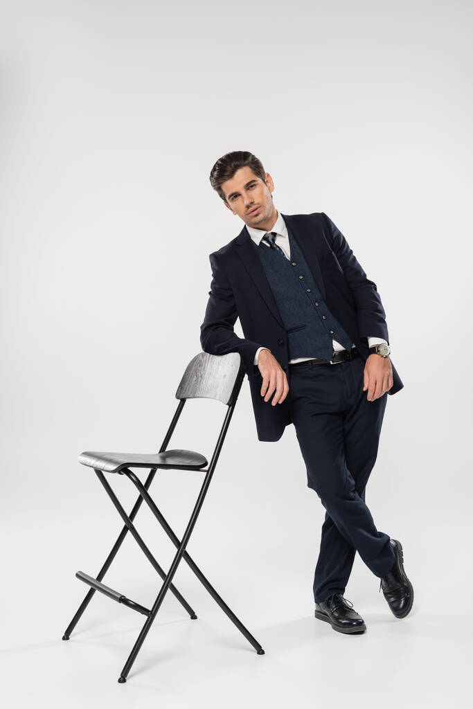piena lunghezza di uomo d'affari di successo in abito con blazer in piedi vicino sedia su grigio - Foto, immagini