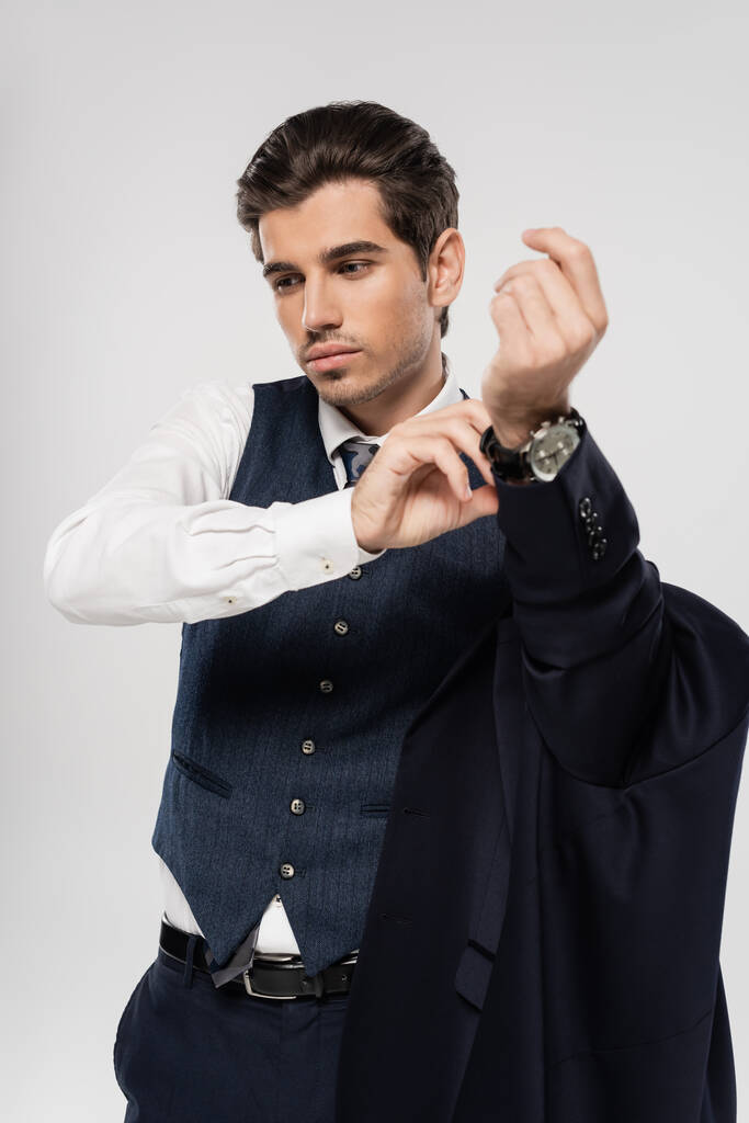 бизнесмен в костюме снимает пиджак и регулирует наручные часы, изолированные на сером - Фото, изображение