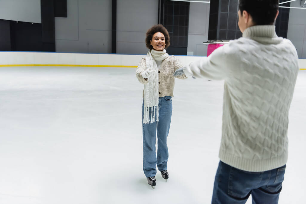 Cheerful african american woman outstretching hands to blurred boyfriend on ice rink  - Photo, Image