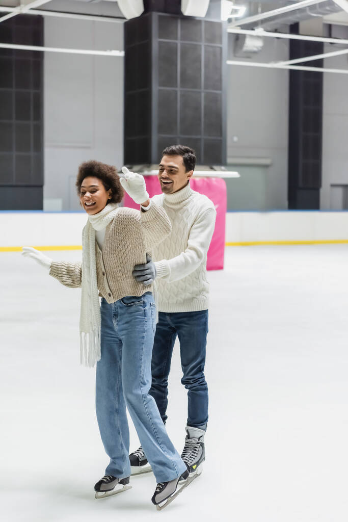 Young man in sweater hugging african american girlfriend skating on ice rink  - Photo, Image