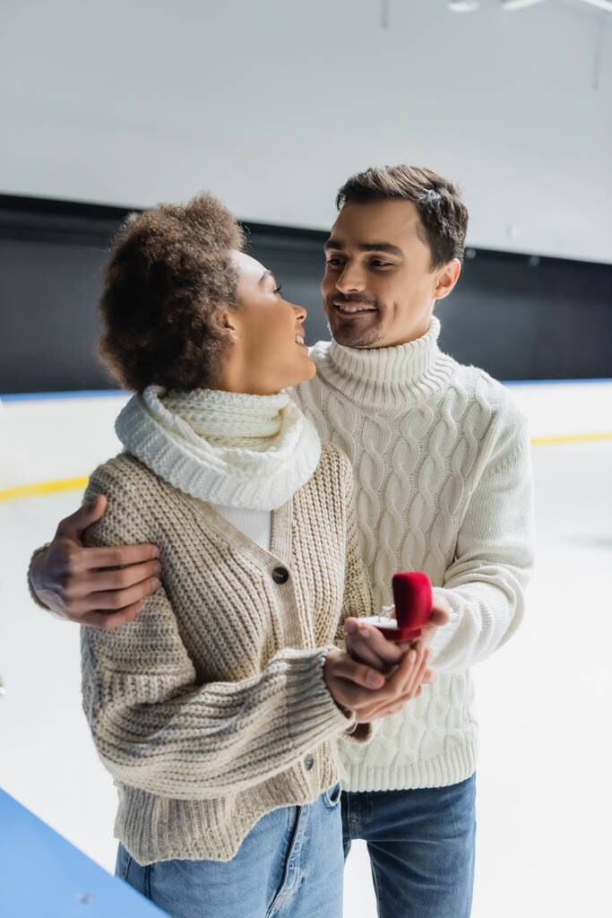 Sonriente mujer afroamericana mirando a novio con anillo de propuesta borrosa en pista de hielo  - Foto, Imagen