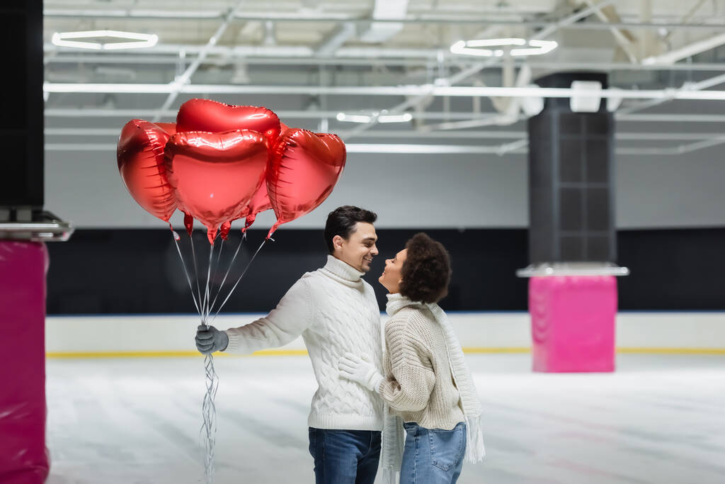 Zijaanzicht van glimlachende Afro-Amerikaanse vrouw knuffelen vriendje met ballonnen in de vorm van hart op de ijsbaan  - Foto, afbeelding