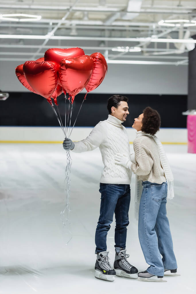 Widok z boku uśmiechnięty mężczyzna trzyma balony w kształcie serca w pobliżu Afryki amerykańskiej dziewczyny na lodowisku  - Zdjęcie, obraz