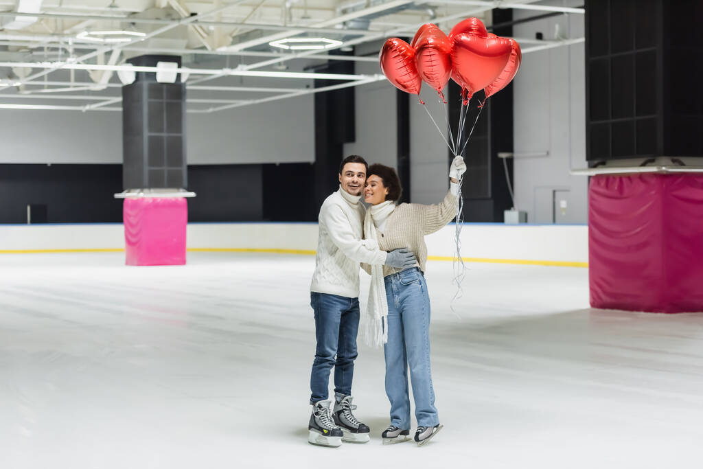Positivo casal interracial abraçando e segurando balões em forma de coração na pista de gelo  - Foto, Imagem