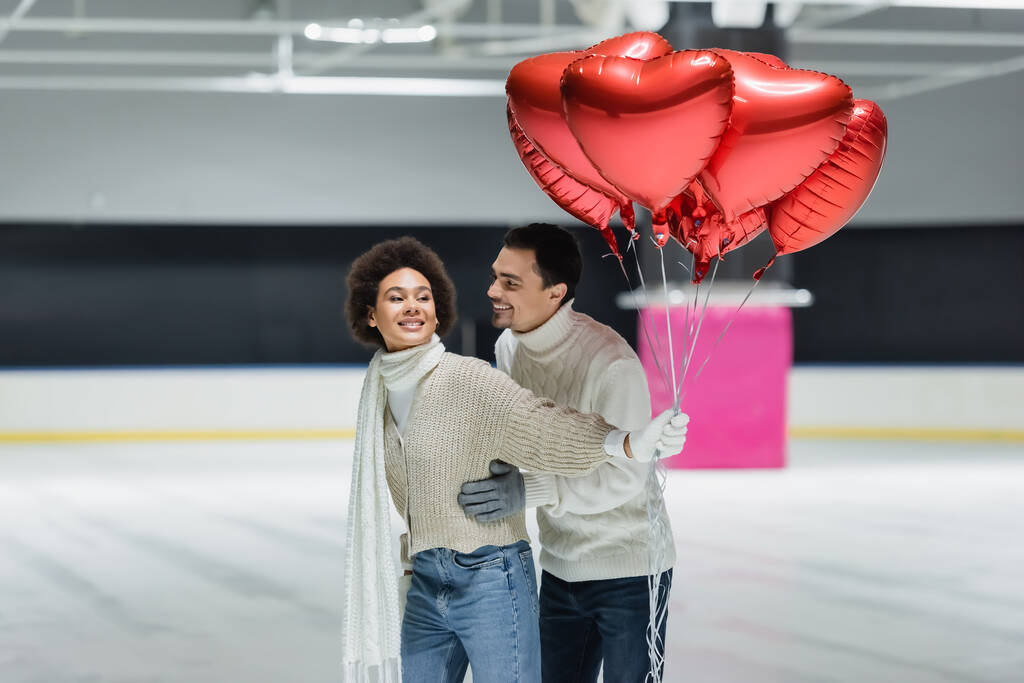 Smiling man in sweater hugging african american girlfriend with balloons in heart shape on ice rink  - Photo, Image
