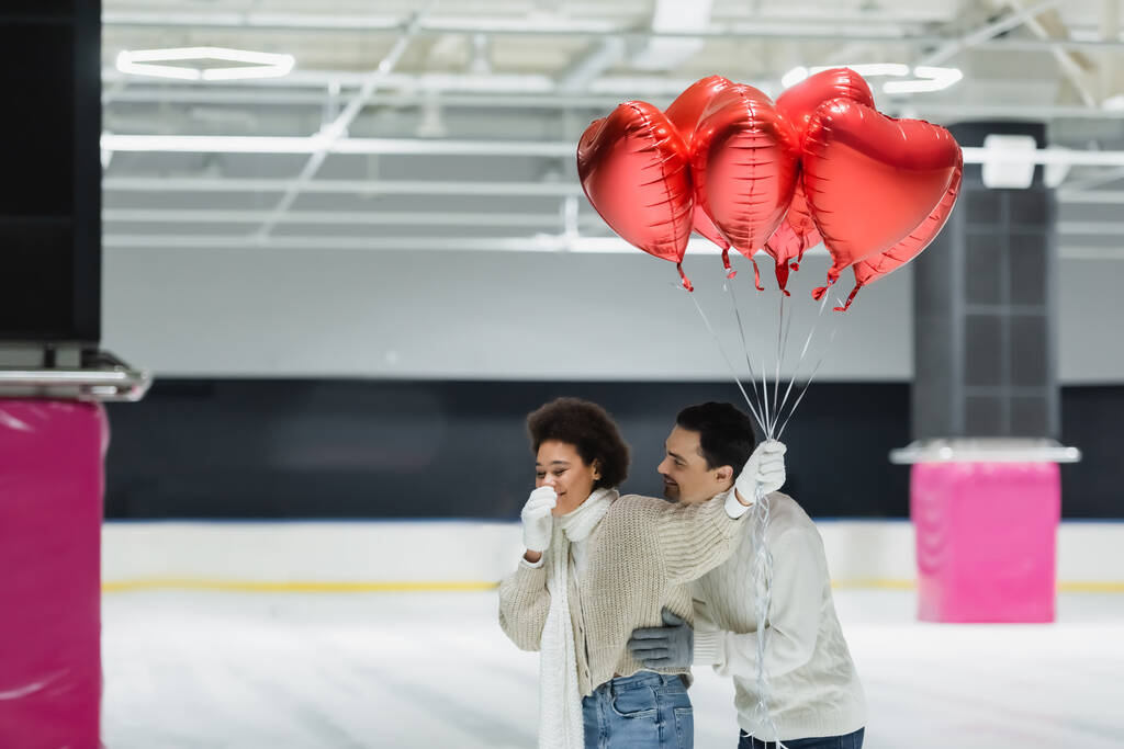 Jovem abraçando alegre afro-americana namorada com balões em forma de coração na pista de gelo  - Foto, Imagem