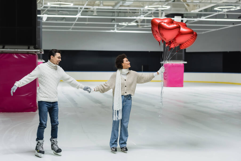 Вид збоку усміхненої міжрасової пари, що тримає повітряні кулі у формі серця під час катання на ковзанах
  - Фото, зображення