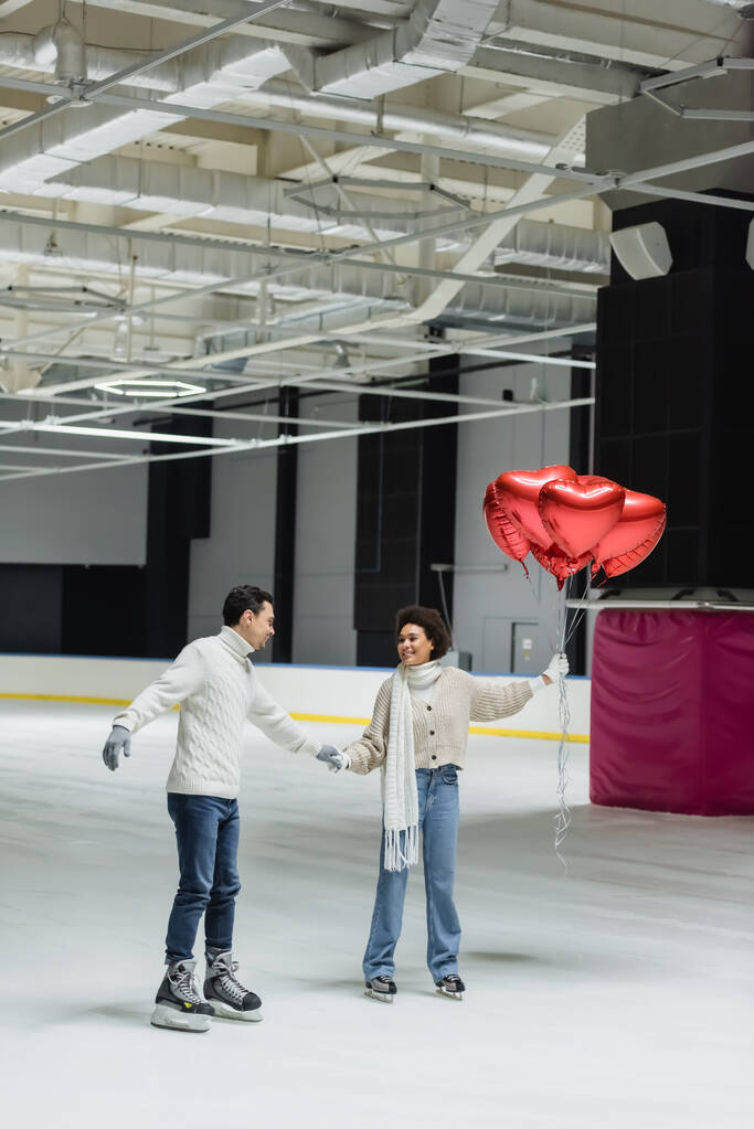 Sorrindo afro-americana segurando balões em forma de coração e mão de namorado na pista de gelo  - Foto, Imagem
