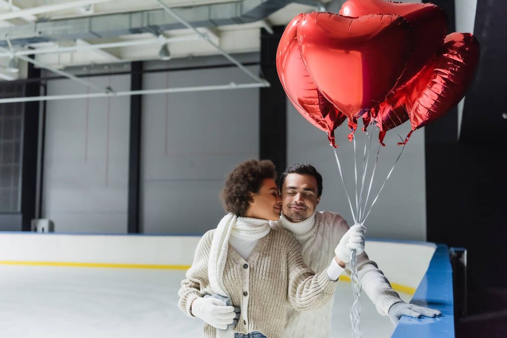 Afrika kökenli Amerikalı bir kadın erkek arkadaşını öpüyor ve buz pistinde kalp şeklinde balonlar tutuyor.  - Fotoğraf, Görsel