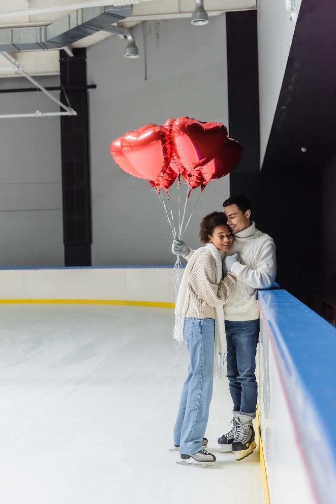 Wesoła międzyrasowa para trzymająca się za ręce i balony w kształcie serca na lodowisku  - Zdjęcie, obraz
