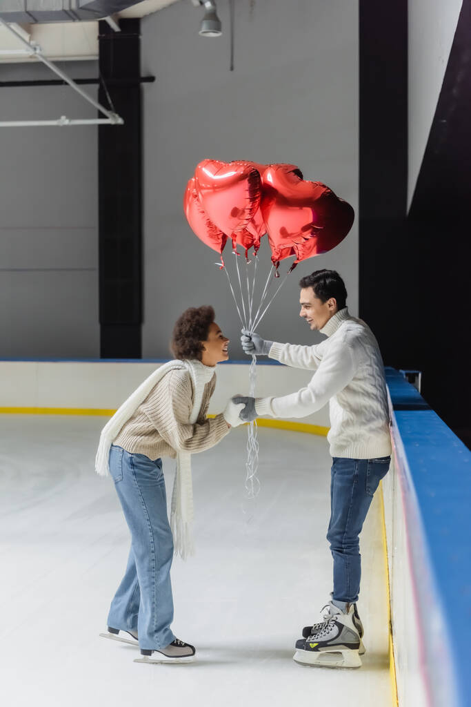 Vista lateral de una mujer afroamericana positiva cogida de la mano de un novio con globos en forma de corazón en una pista de hielo  - Foto, imagen
