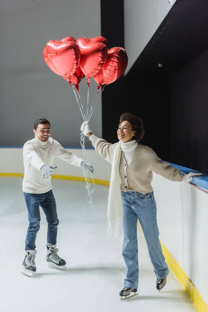 Sonriente mujer afroamericana sosteniendo globos rojos en forma de corazón mientras patina sobre hielo cerca de su novio en la pista  - Foto, imagen