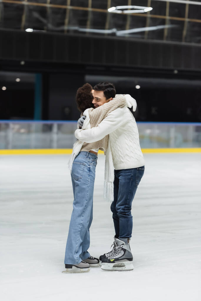 Hombre sonriente en suéter abrazando novia afroamericana en pista de hielo  - Foto, Imagen