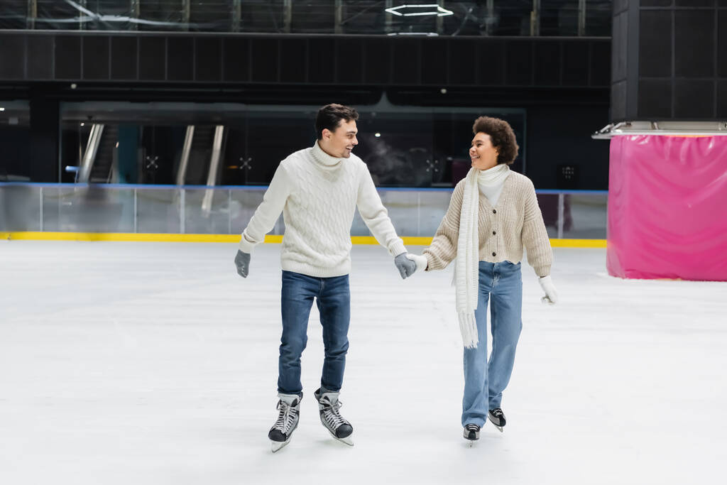 Cheerful interracial couple in gloves and sweaters skating on ice rink  - Photo, Image