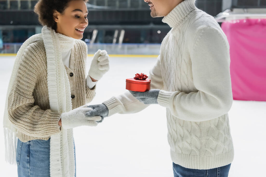 Glimlachende man met hartvormige doos en hand van Afrikaanse Amerikaanse vriendin op de ijsbaan  - Foto, afbeelding