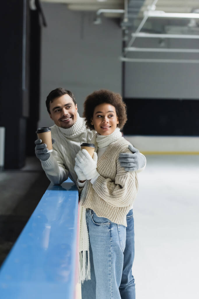 Hombre sonriente abrazando novia afroamericana con café para ir en pista de hielo  - Foto, imagen