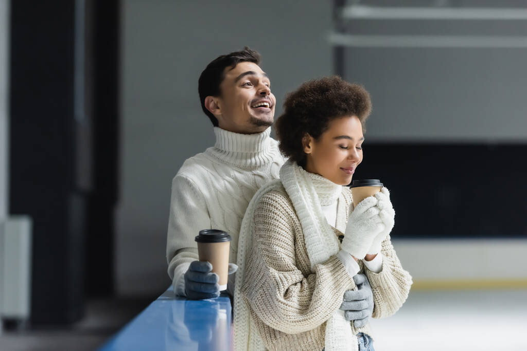 glimlachende man in trui houden koffie te gaan en knuffelen Afrikaans amerikaanse vriendin op de ijsbaan  - Foto, afbeelding