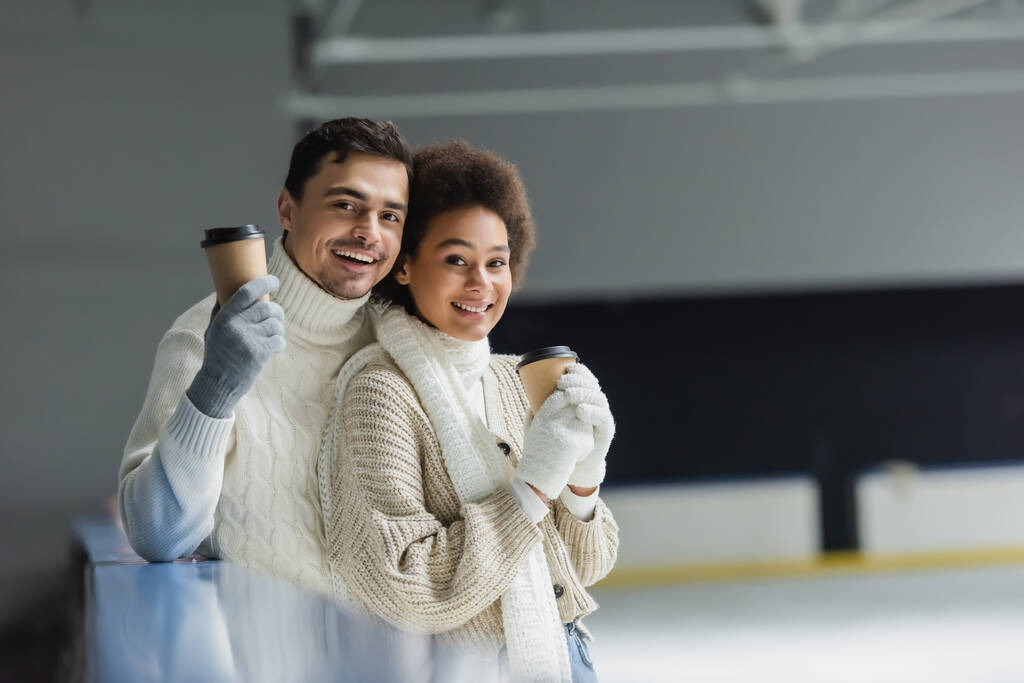 Allegro coppia interrazziale in abiti caldi che tengono il caffè per andare e guardando la fotocamera sulla pista di pattinaggio sul ghiaccio - Foto, immagini