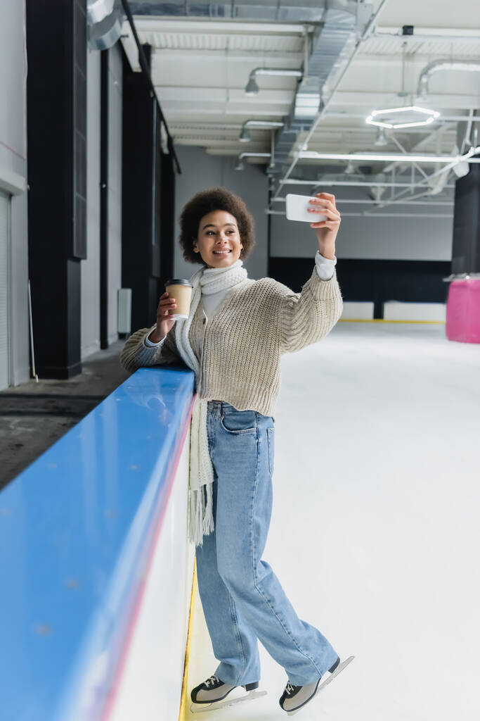 Mujer afroamericana en patines de hielo sosteniendo café para ir y tomar selfie en smartphone en pista de hielo  - Foto, imagen