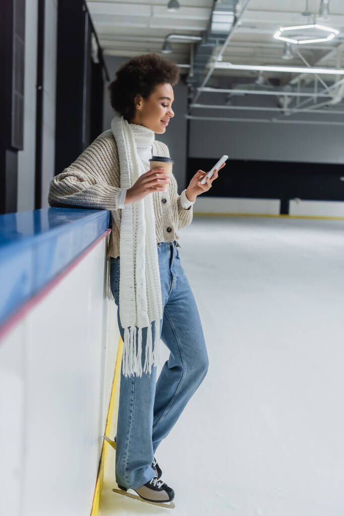 Widok z boku uśmiechniętej Afroamerykanki na łyżwach za pomocą telefonu komórkowego i trzymając kawę na lodowisku  - Zdjęcie, obraz