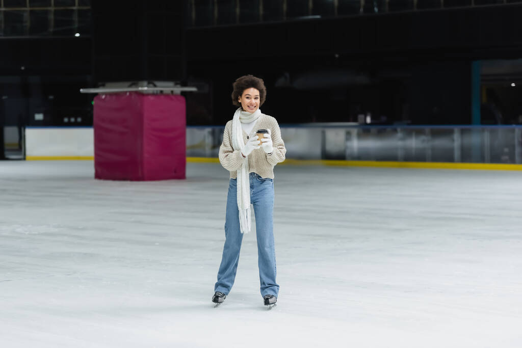 Cheerful african american woman holding paper cup and looking at camera on ice rink  - Photo, Image
