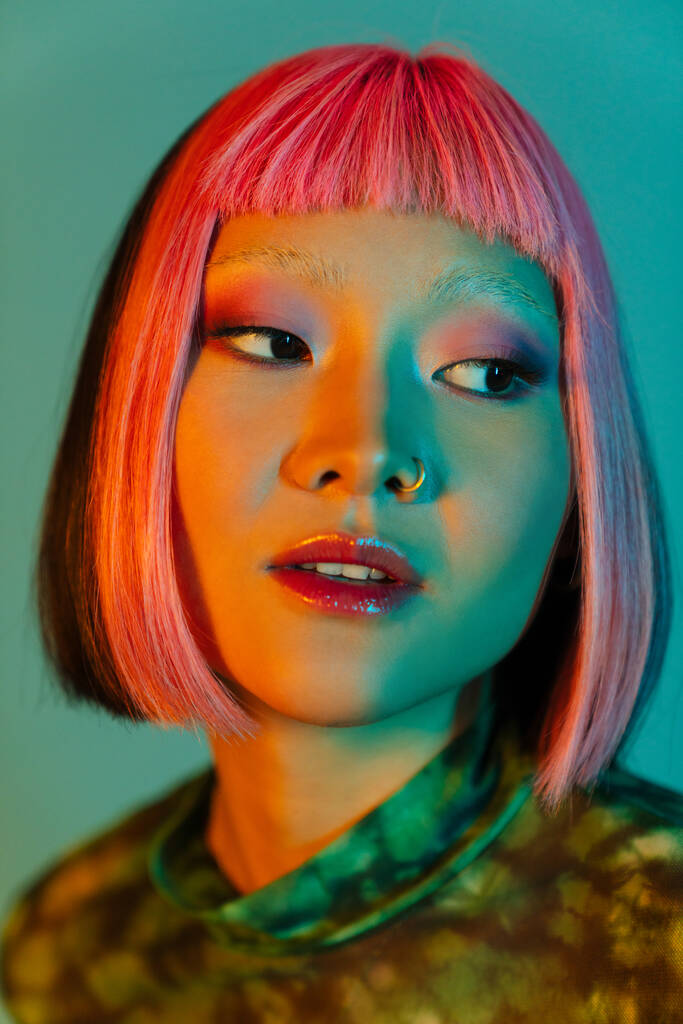Asiatisches Mädchen mit pinkfarbenen Haaren und Piercing posiert, während es isoliert über blauem Hintergrund zur Seite schaut - Foto, Bild