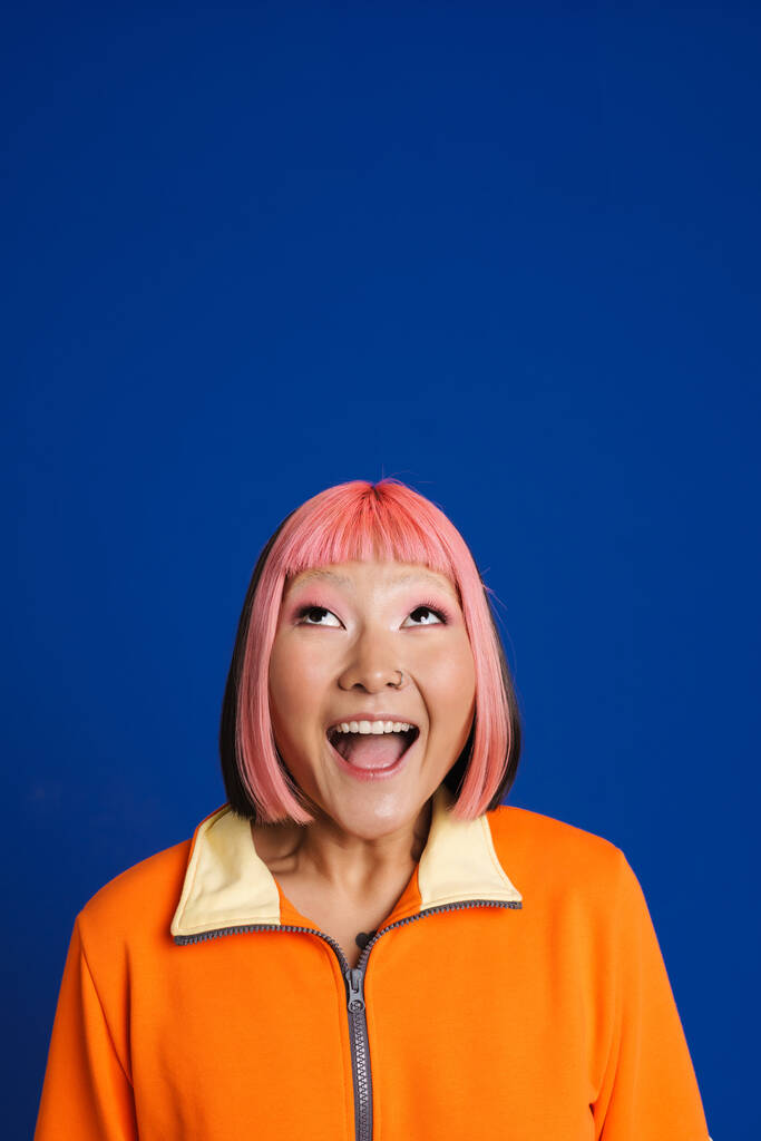 Asiatisches Mädchen mit pinkfarbenen Haaren und Piercing, das seine Überraschung über die isolierte Kamera auf blauem Hintergrund ausdrückt - Foto, Bild