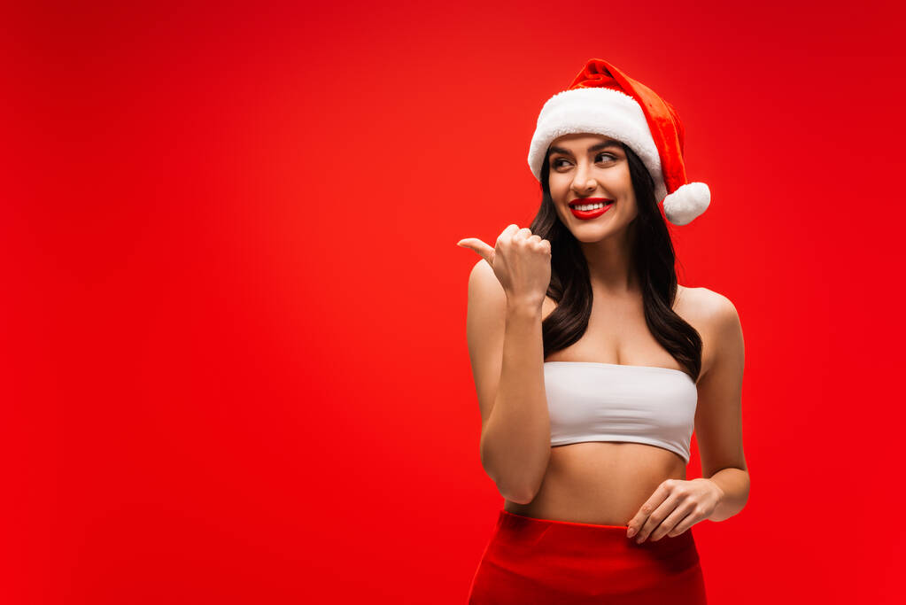 усміхнена жінка в капелюсі Санта, вказуючи пальцем ізольовано на червоному
 - Фото, зображення