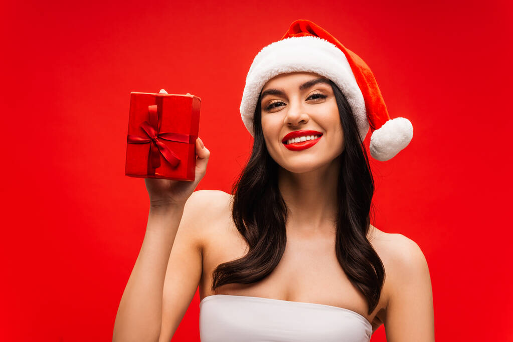Positive brunette model in santa hat holding present isolated on red - Photo, Image