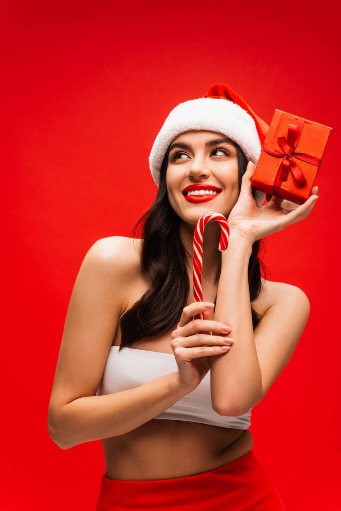 Cheerful young woman in santa hat holding striped lollipop and present isolated on red - Photo, Image