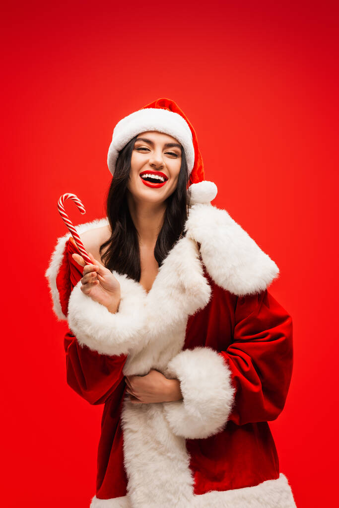 Veselý mladý model v kostýmu Santa drží pruhované lízátko izolované na červené - Fotografie, Obrázek