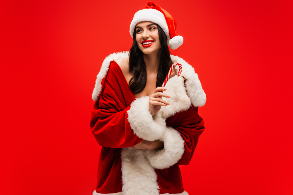 Positive brunette model in santa costume holding striped lollipop isolated on red - Photo, Image