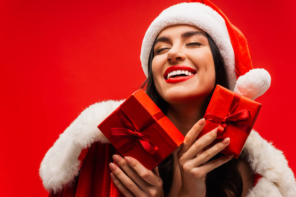 Glückliche Frau mit Weihnachtsmannhut hält Geschenkschachteln in der Hand und schließt die Augen isoliert auf Rot - Foto, Bild