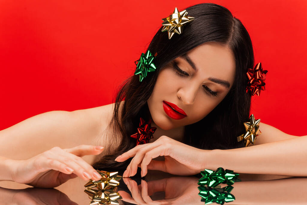 Красивая женщина с обнаженными плечами и блестящими луками подарка касательно отражающей поверхности, изолированной на красном  - Фото, изображение