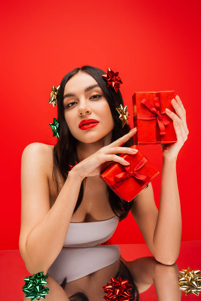 Jeune modèle en haut et noeuds cadeaux sur la tenue de cheveux présente près de surface réfléchissante isolé sur rouge  - Photo, image