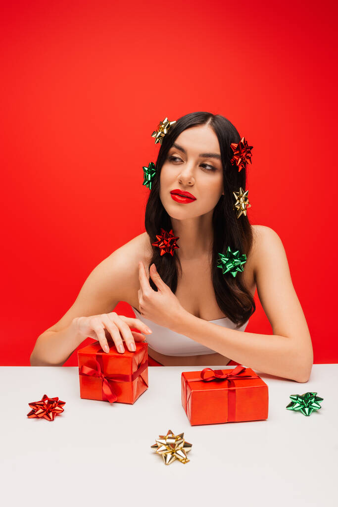 Mujer joven bonita con lazos de regalo en el cabello mirando hacia otro lado cerca de regalos aislados en rojo  - Foto, Imagen