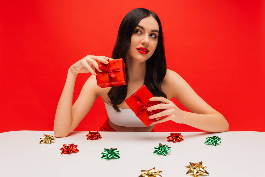 Femme brune avec maquillage tenant des cadeaux près de noeuds cadeaux brillants isolés sur rouge  - Photo, image