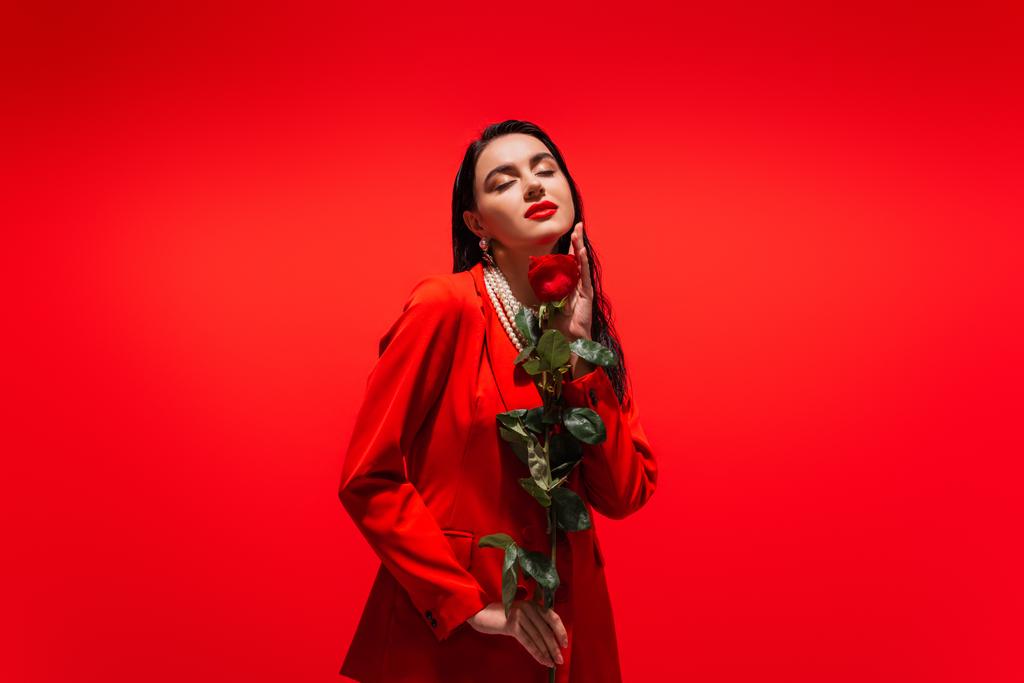 Όμορφη μελαχρινή γυναίκα με σακάκι που κρατά το τριαντάφυλλο απομονωμένο στο κόκκινο  - Φωτογραφία, εικόνα