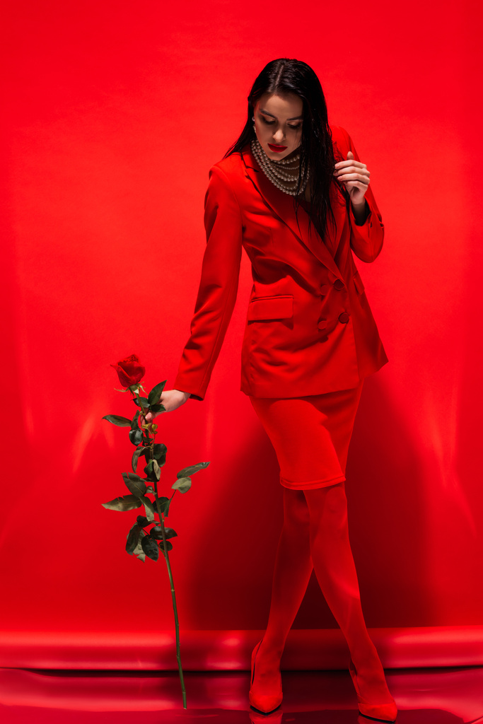 Volledige lengte van stijlvolle vrouw in jas en parel ketting houden roos op rode achtergrond  - Foto, afbeelding