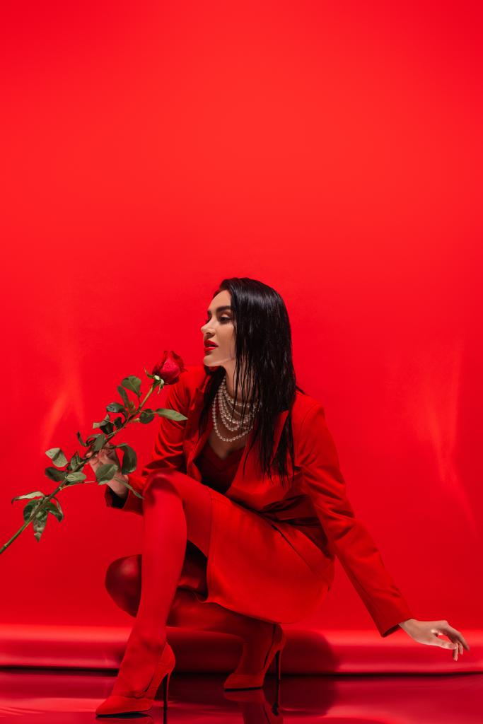 Стильна жінка на підборах і перловому намисті тримає троянду на червоному тлі
  - Фото, зображення
