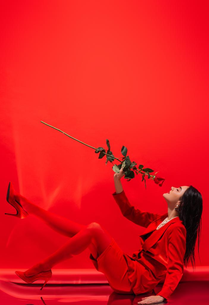Вид сбоку на брюнетку модели в каблуках, пахнущих розой, сидя на красном фоне  - Фото, изображение
