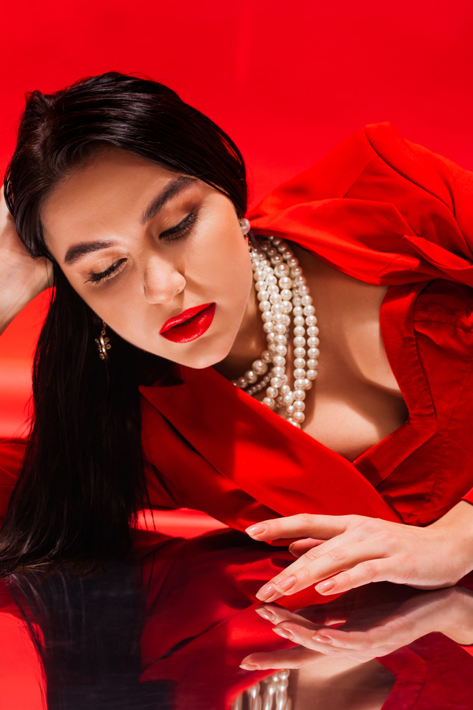 Stilvolle brünette Frau in Perlenkette berühren reflektierende Oberfläche auf rotem Hintergrund  - Foto, Bild