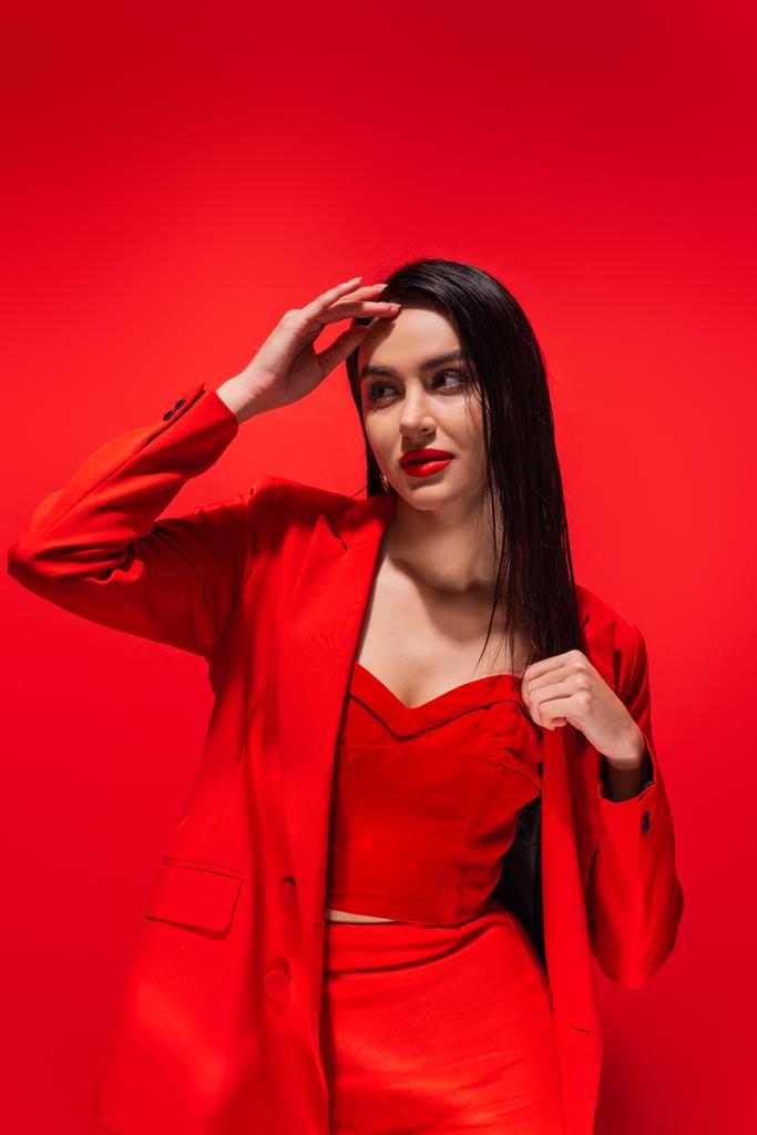 Stylish brunette woman in jacket posing and looking away isolated on red  - Photo, Image