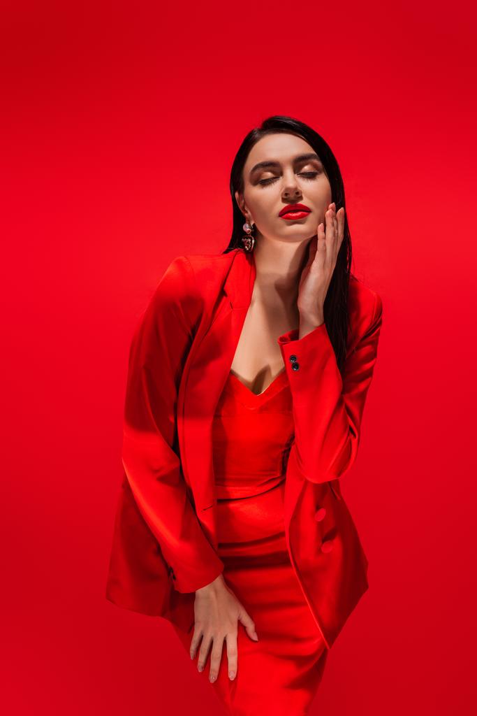 Modelo elegante e elegante na jaqueta tocando o rosto e fechando os olhos isolados no vermelho  - Foto, Imagem