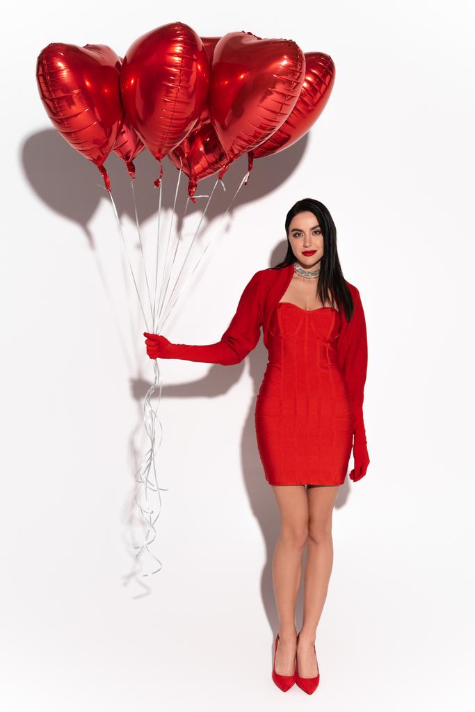 Volle Länge der stilvollen Frau in rotem Kleid und High Heels mit herzförmigen Luftballons auf weißem Hintergrund mit Schatten  - Foto, Bild