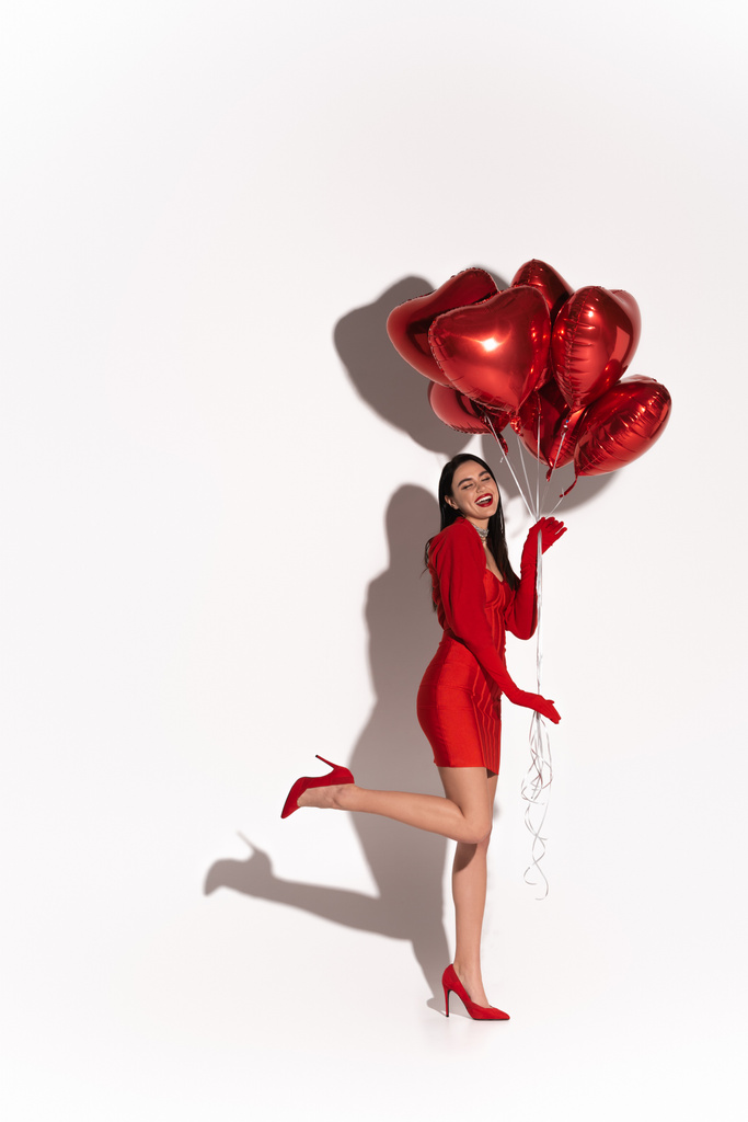 Longitud completa de mujer bonita en tacones rojos y vestido posando con globos en forma de corazón sobre fondo blanco con sombra  - Foto, Imagen