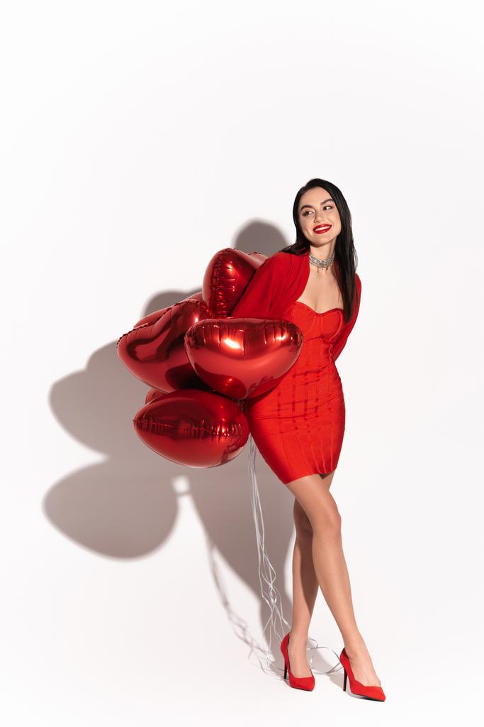 Стильная брюнетка женщина на каблуках глядя в сторону красных шаров в форме сердца на белом фоне с тенью  - Фото, изображение