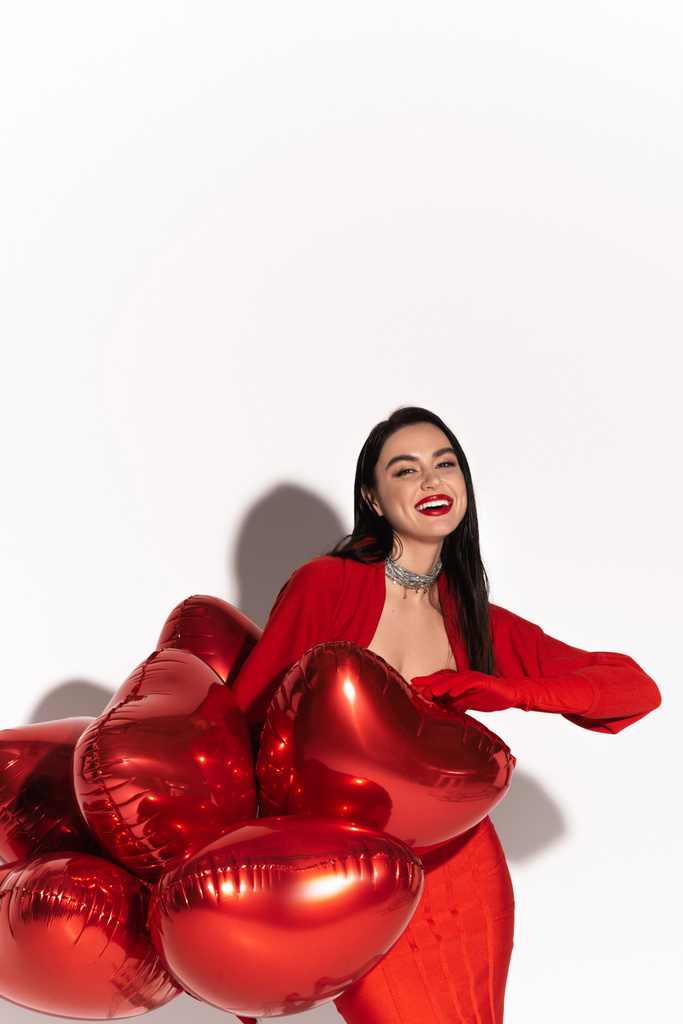 Stylish woman with red lips looking at camera near heart shaped balloons on white background with shadow  - Photo, Image