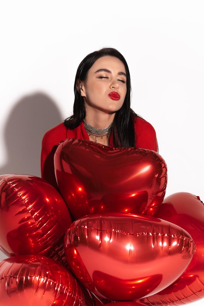 Mujer con estilo con labios rojos soplando beso cerca de globos en forma de corazón sobre fondo blanco con sombra  - Foto, Imagen