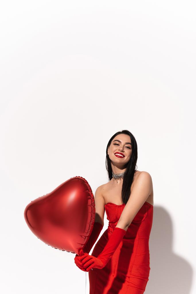 Mulher sorridente em vestido vermelho segurando balões em forma de coração e olhando para a câmera no fundo branco com sombra  - Foto, Imagem